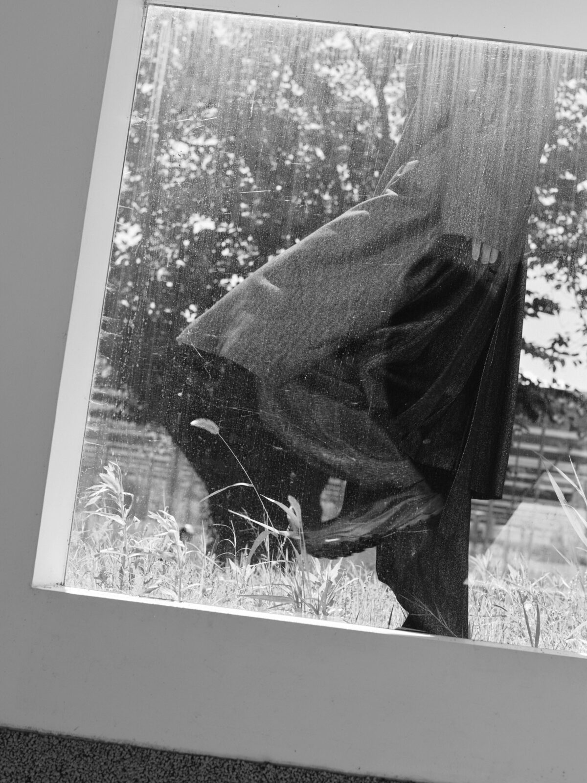 草木を映す窓越しに、黒い服とシューズを身につけた人物が重なるモノクロ写真